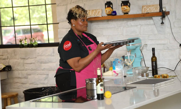 A woman wearing a black t-shirt and a bright pink apron stands in a kitchen setting, holding a metal tray. The apron features a red circular logo on the left chest that reads 'food'.