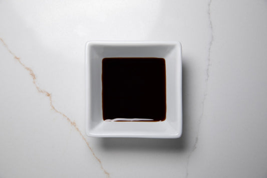 A square white ceramic dish containing a dark brown liquid, likely soy sauce, set on a light gray marble surface.