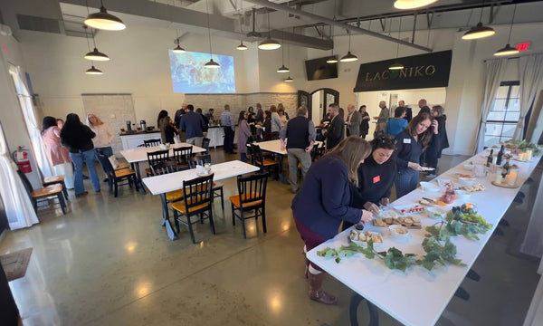 A bustling indoor event space with many people mingling around tables and a long serving table in the foreground. The room has exposed ceiling details, hanging pendant lights, a concrete floor, and a black wall sign reading LAZARO.