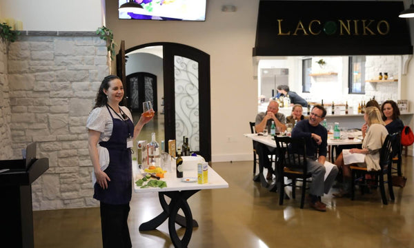 A woman in a dark navy apron over a white shirt stands in a restaurant holding a wine glass, with a table of wine bottles in front of her and a group of people dining in the background under a black 'LACONIKO' sign.