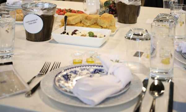 A close-up of a formal dining table setting with a blue and white patterned plate, white napkin, silver cutlery, clear drinking glasses, and a white tablecloth. In the background, a white rectangular dish holds small appetizers, and other tableware is visible.