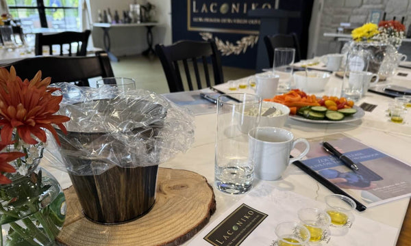 A dining table set in a restaurant or event space, featuring a dark wood bucket wrapped in clear plastic, white mugs, water glasses, a plate of sliced cucumbers and tomatoes, and a Lagoniko sign in the background.