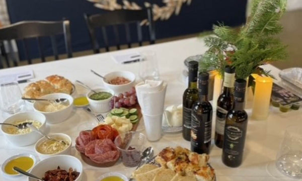 A styled dining table laden with an assortment of food items, including bowls of pasta, cured meats, sliced tomatoes and cucumbers, bottles of olive oil and wine, and candles, with a greenery centerpiece.