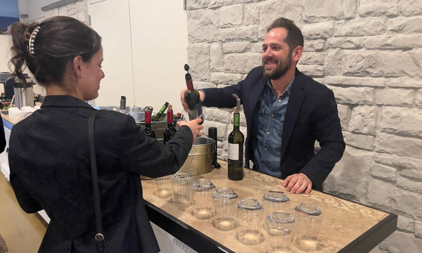 A man in a navy blazer and blue shirt pours wine from a bottle into a glass held by a woman wearing a black blazer, set against a white brick wall background.