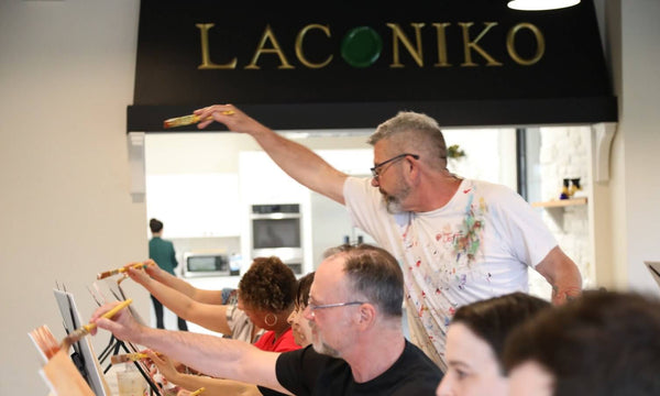 A group of people participating in a painting workshop or class, with one man standing and gesturing while others paint at easels under a black sign that reads 'LACONIKO'.