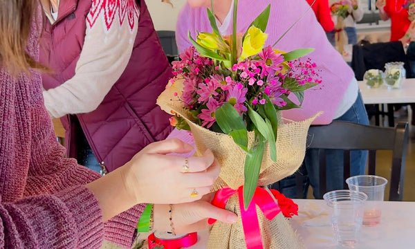 A close-up of a person holding a small bouquet of pink and yellow flowers wrapped in burlap and tied with a bright pink ribbon, in a social indoor setting with other people and tables visible in the background.